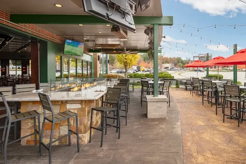 Outdoor sports bar patio with fire pit, TVs, and red umbrellas.