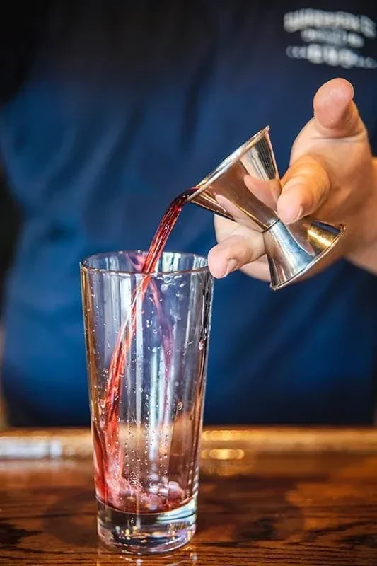A hand pours red liquid from a silver jigger into a clear glass.