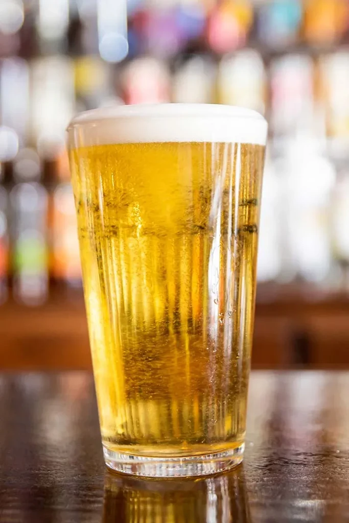Glass of golden beer with foam and condensation, blurry background.
