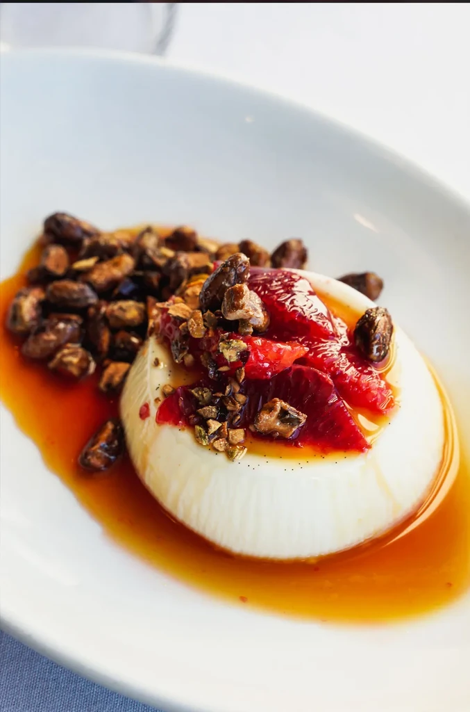 Molded dessert with red fruit, nuts, and amber syrup in a white dish.