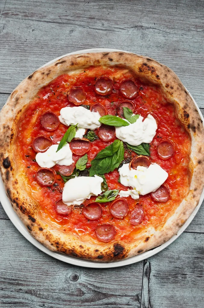 Overhead Neapolitan pepperoni pizza with cheese, basil on a wooden table.