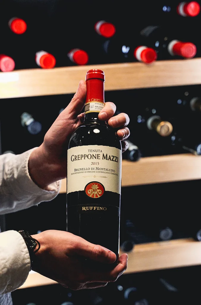 Two hands present a Tenuta Greppone Mazzi Brunello di Montalcino 2015 bottle.