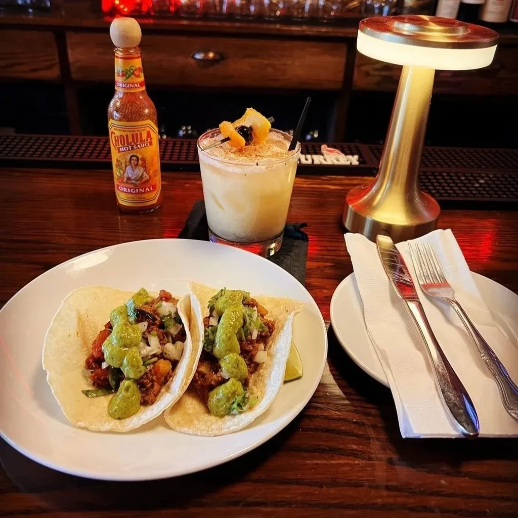Two tacos, a cocktail, and Cholula hot sauce on a wooden bar.