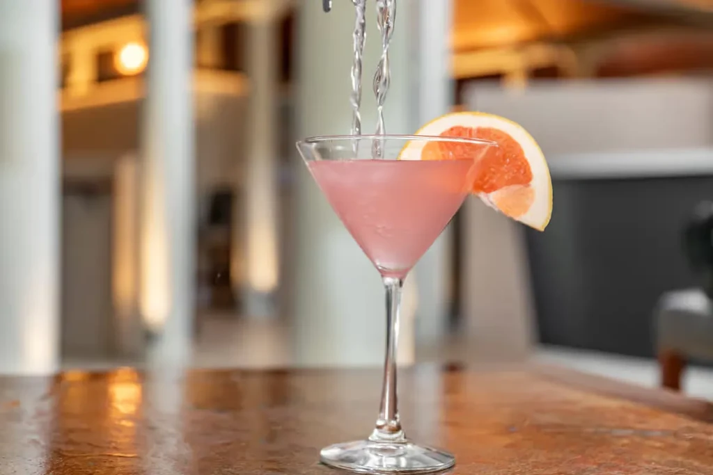 Pink cocktail in martini glass, grapefruit garnish, liquid pouring.