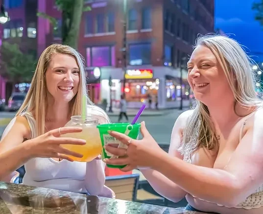 Two blonde women clink drinks and laugh in an outdoor city setting.