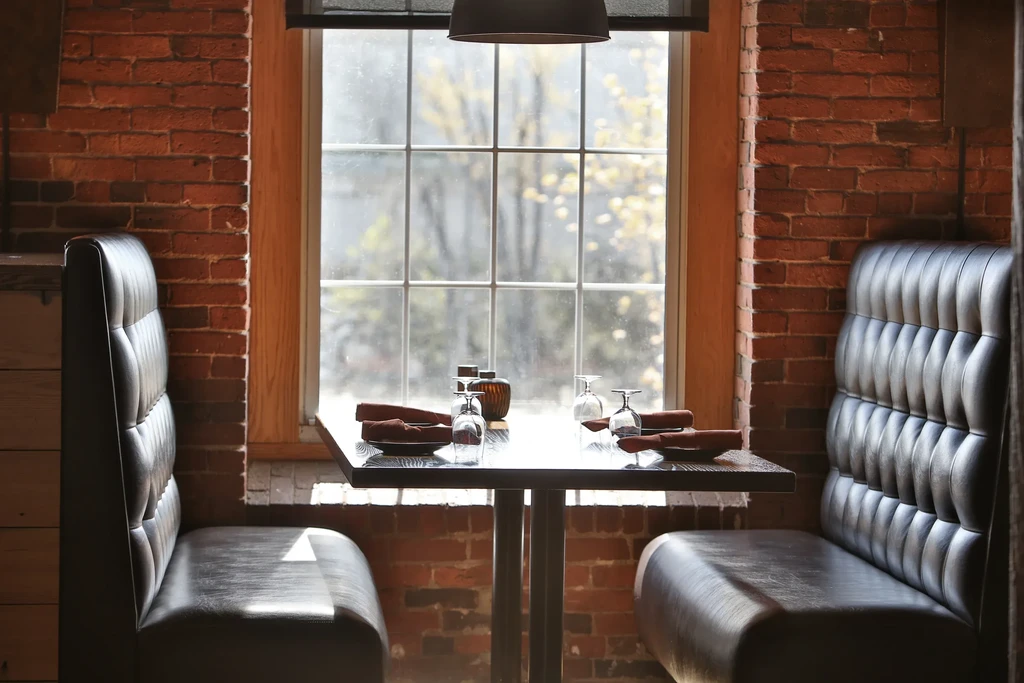Cozy restaurant booth with tufted benches, brick walls, window, set table.