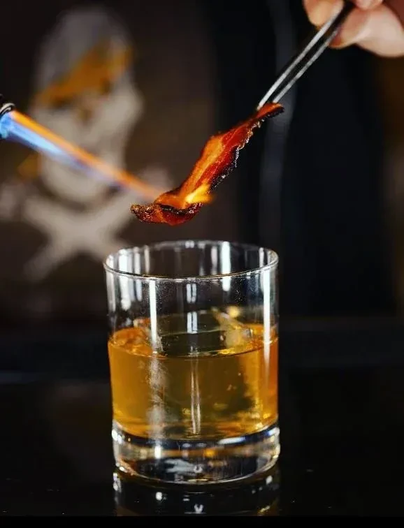 Bacon strip torched over amber cocktail with ice; skull in background.