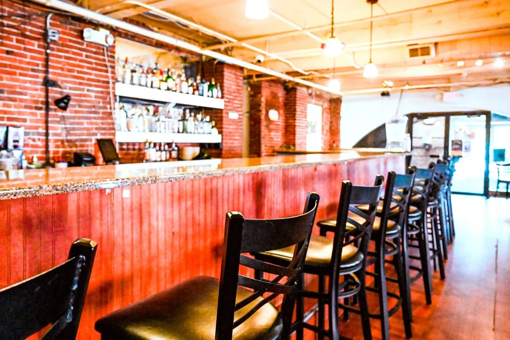 Rustic bar interior with wooden counter, brick wall, bottles, and stools.