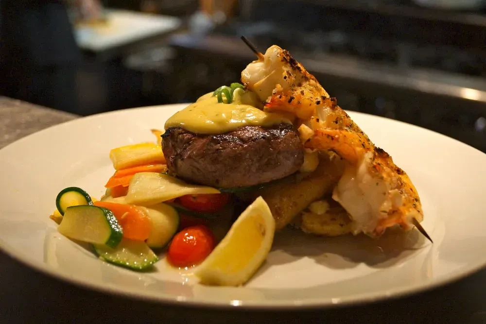 Plate with steak, grilled seafood skewer, colorful vegetables, and lemon.