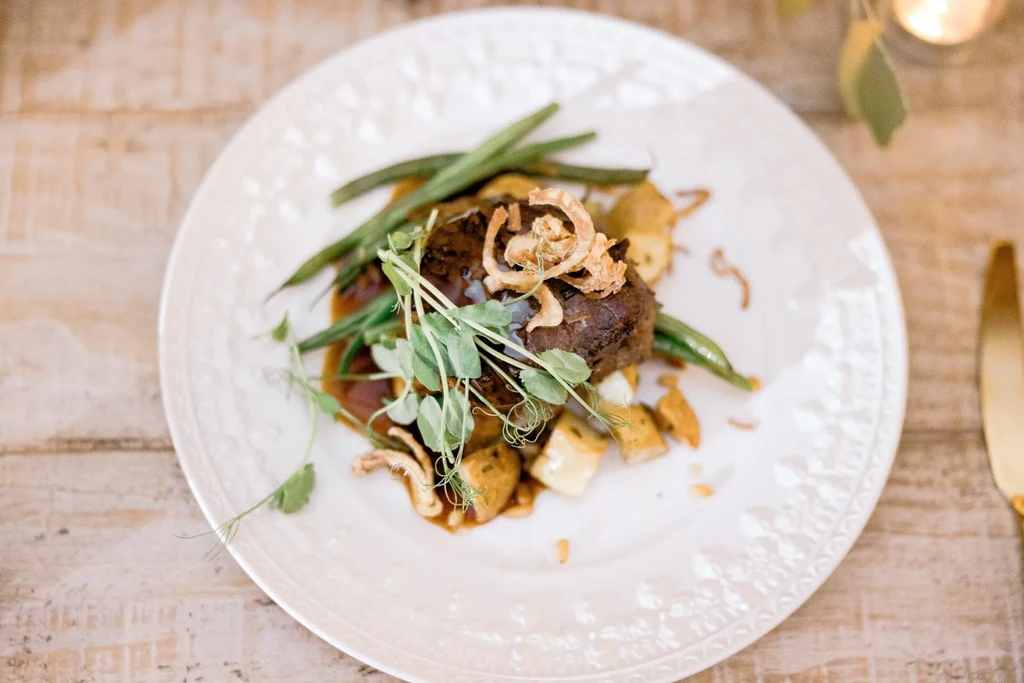 Overhead view of meat entree with sauce, pea shoots, green beans, potatoes.