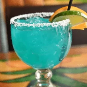 Turquoise drink in goblet, salted rim, crushed ice, lime, orange garnish.
