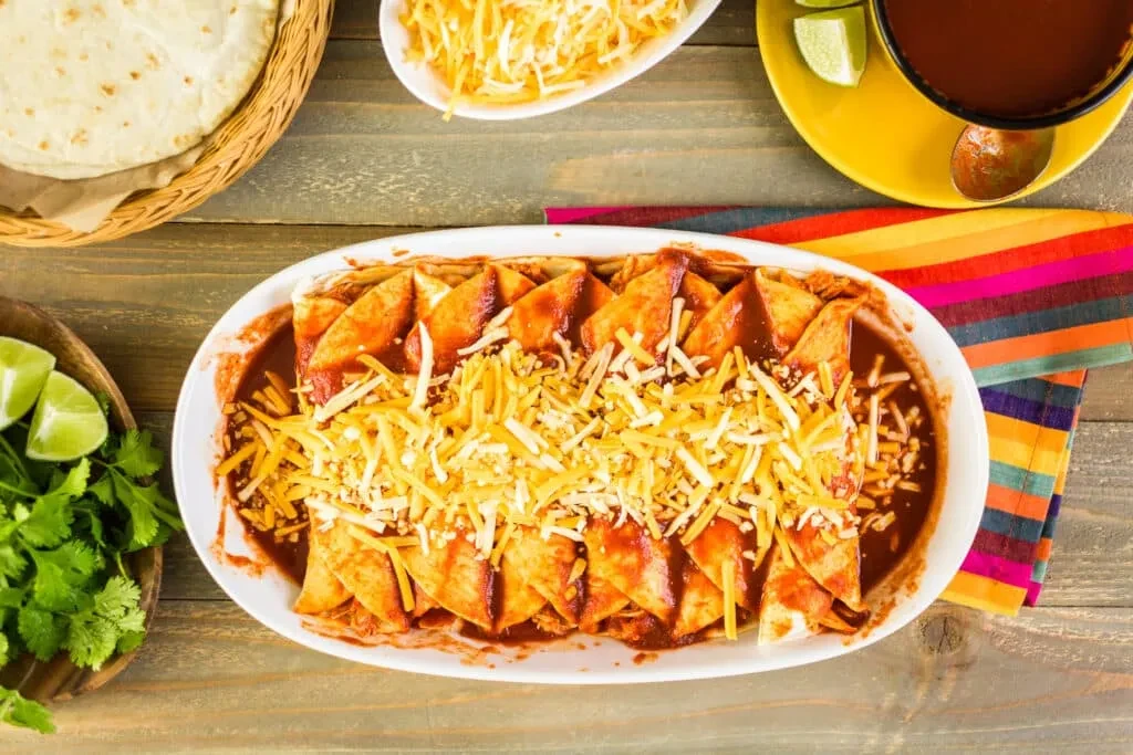 Top-down view: Saucy enchiladas with cheese, tortillas, cilantro, limes on wood.