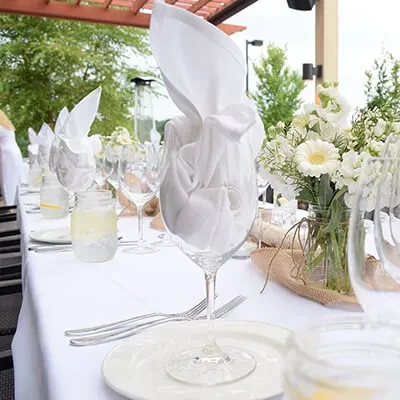 Neatly set outdoor dining table with white place settings, flowers, drinks.