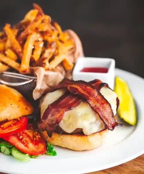 Burger with melted cheese, bacon, lettuce, tomato, fries, ketchup, and pickle.