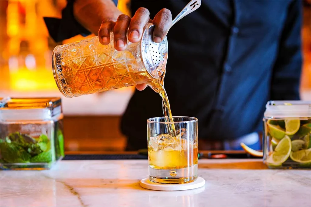 Close-up: dark hand strains amber drink into ice-filled glass, fresh ingredients nearby.