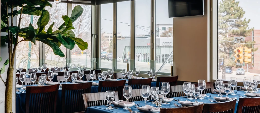 Empty restaurant dining room features set tables, large plant, and city view.
