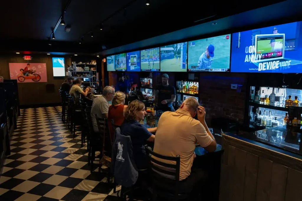 Dimly lit bar with patrons at counter, TVs, and checkerboard floor.