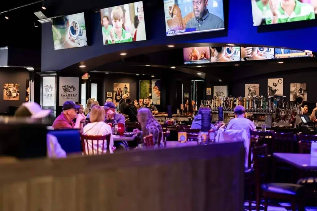 Lively purple-lit bar interior with many patrons, TVs, and stocked bar.