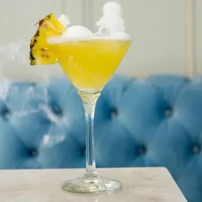 Vibrant yellow cocktail in martini glass with pineapple, cream, dry ice.