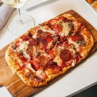 Rectangular pizza with cheese, pepperoni, sausage, and herbs on a wooden board.