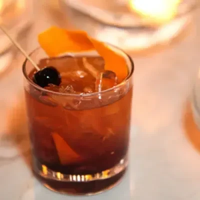 Close-up of an amber cocktail with ice, orange peel, and dark cherry.