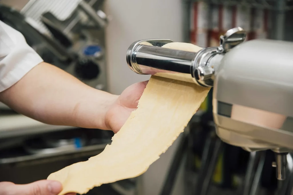 Hands feeding pasta dough through a stand mixer pasta attachment.