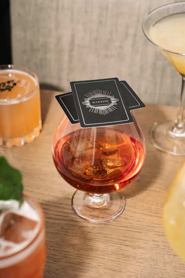 Close-up: Amber snifter cocktail with DANTON cards on its rim.