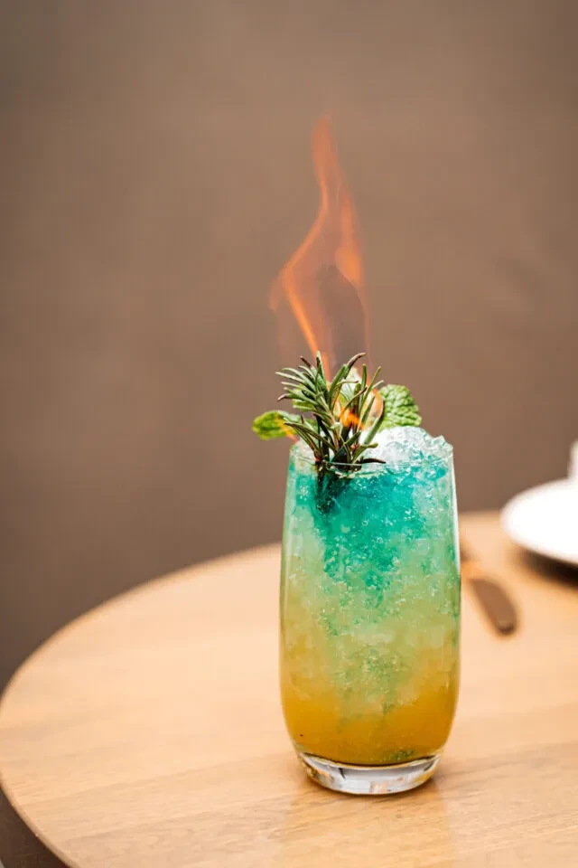 Layered yellow and teal cocktail with crushed ice, burning rosemary, and mint.