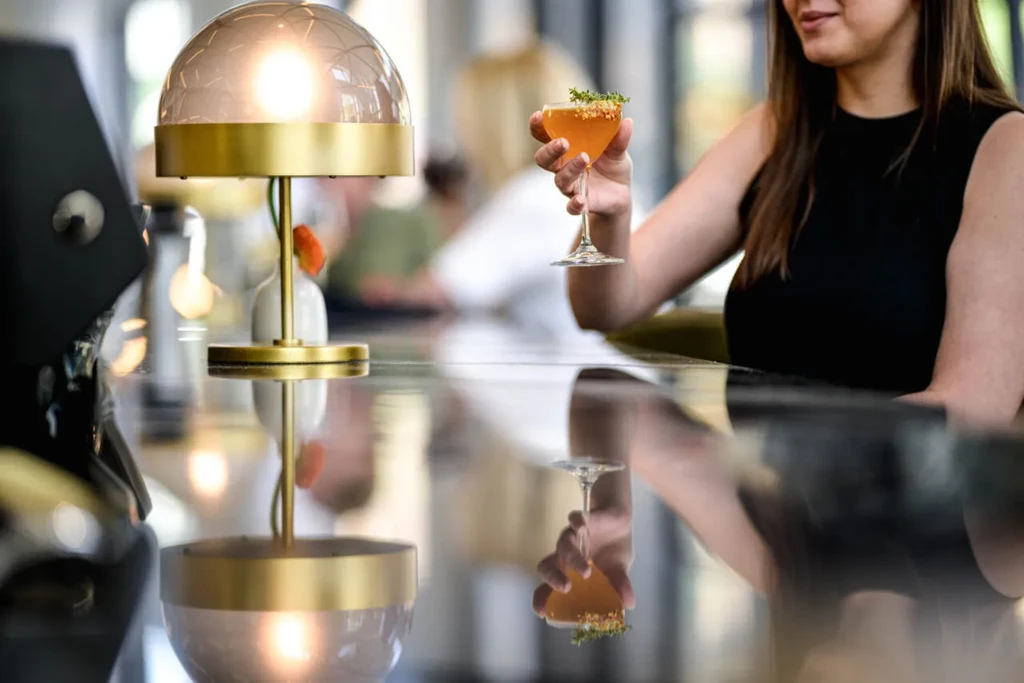 Woman holds garnished amber cocktail, lamp reflects on bar surface.