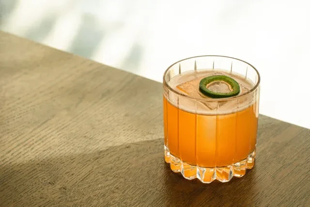 Orange cocktail with frothy top, jalapeño, textured glass on wood.