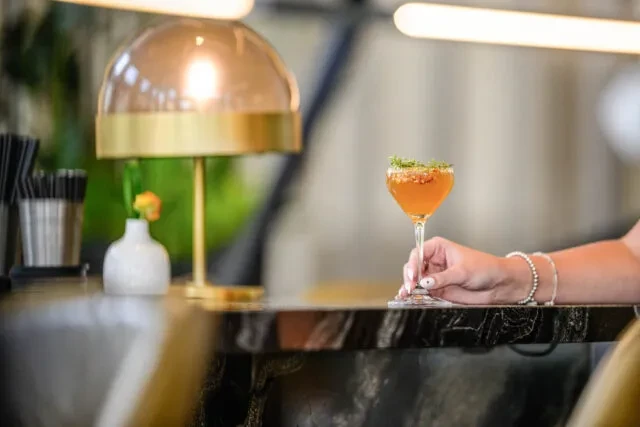 Amber cocktail with green garnish, held by a hand at a bar.