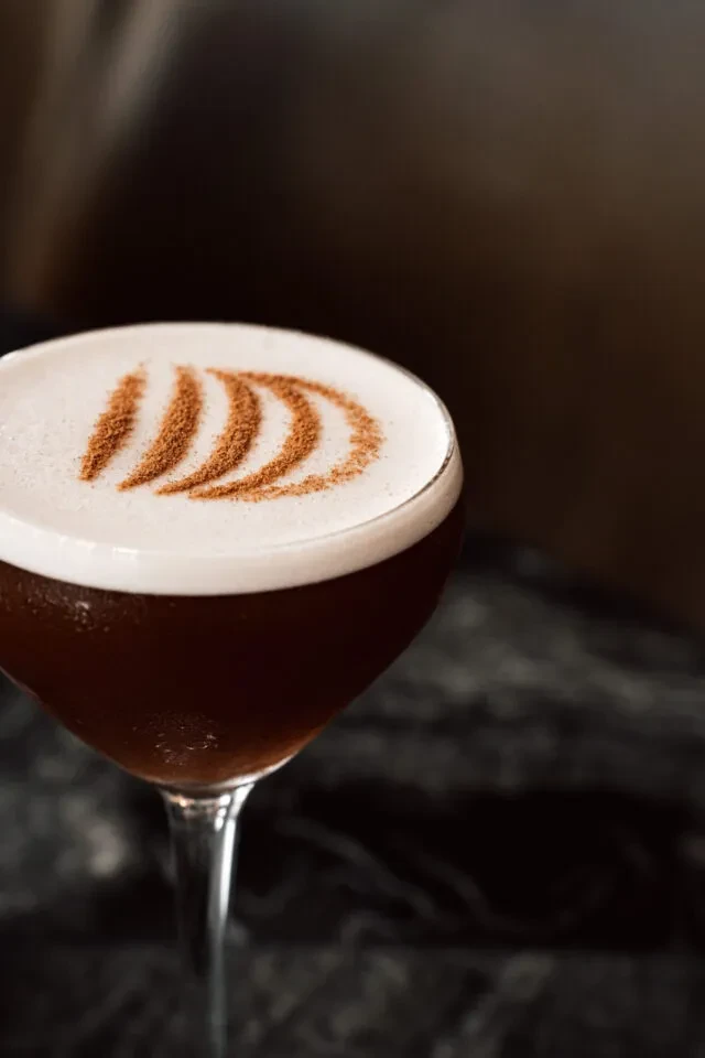 Dark brown cocktail with white foam and powder garnish in a coupe glass.