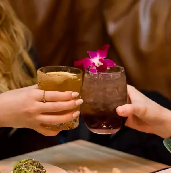 Two hands clink amber cocktail with sugared rim, purple with orchid.