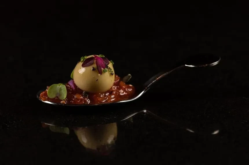 Spherical appetizer with relish, greens, on a silver spoon, black background.
