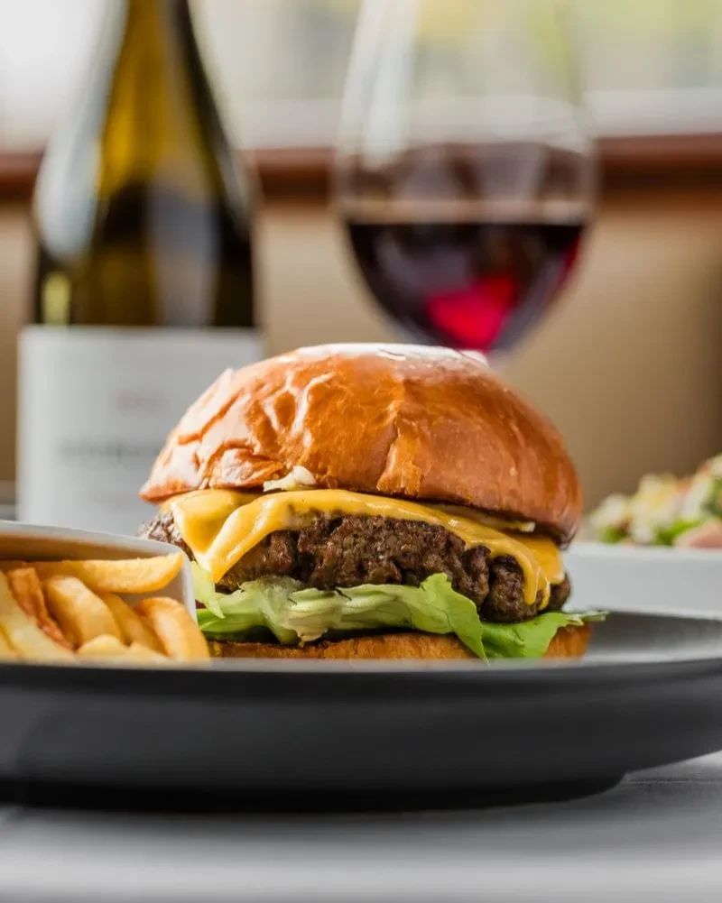 Close-up cheeseburger with melted cheese, fries, and blurry wine background.