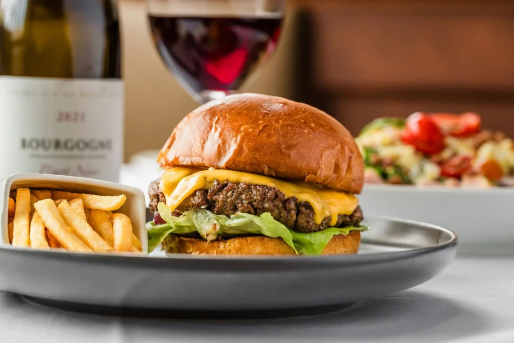 Close-up cheeseburger and crispy fries, with blurred wine background.