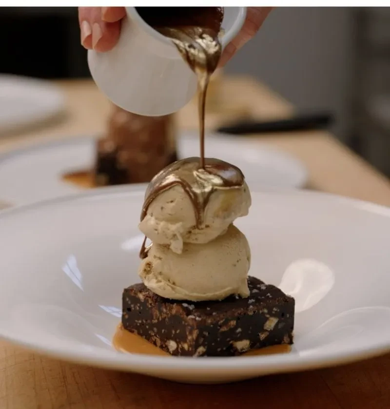 Hand drizzles chocolate sauce over brownie and vanilla ice cream.