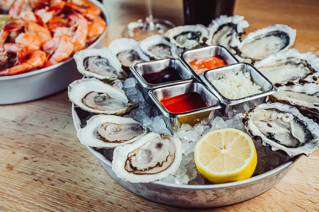 Fresh oysters on ice with lemon and sauces, shrimp in background.