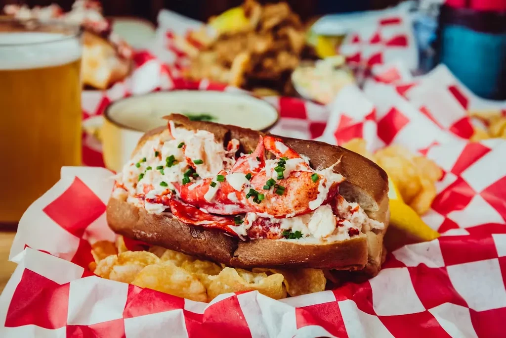 Close-up of a lobster roll, chives, chips, and a glass of beer.