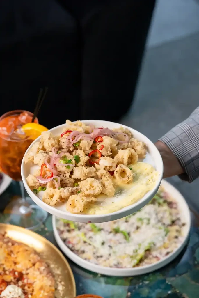Hand holds fried calamari with red onion, chili, herbs, next to cocktail.
