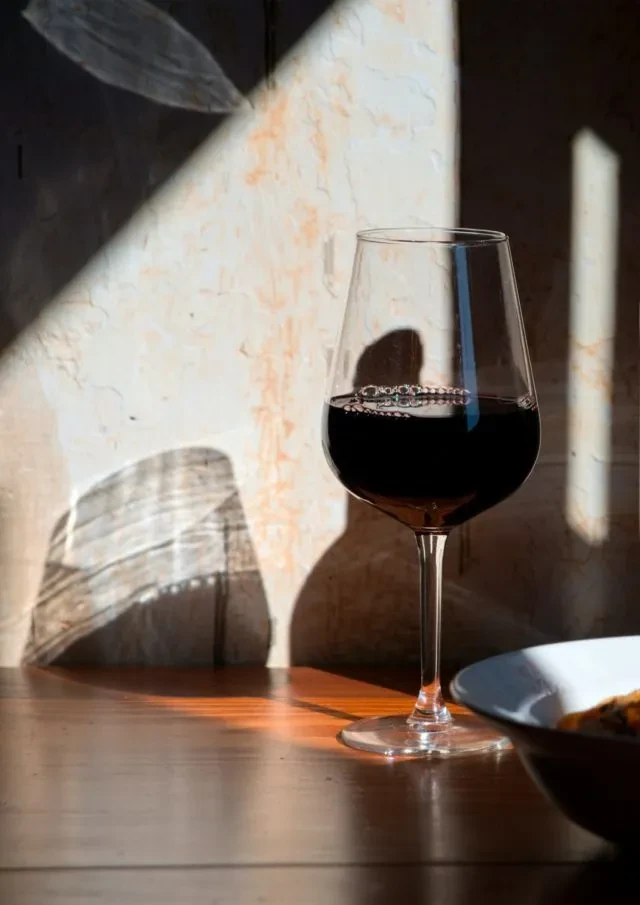 Half-filled red wine glass on wooden table, strong sunlight, shadows.