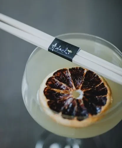 Cocktail with dried orange slice and Adachi-branded decorative sticks.