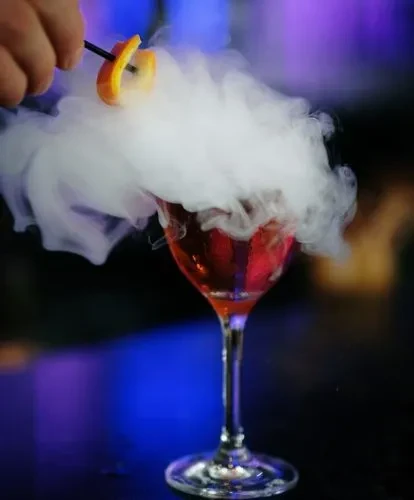Red cocktail in stemmed glass, rising smoke, hand with orange garnish.