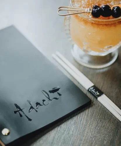 Black 'Adachi' menu, white chopsticks, and amber cocktail on a table.