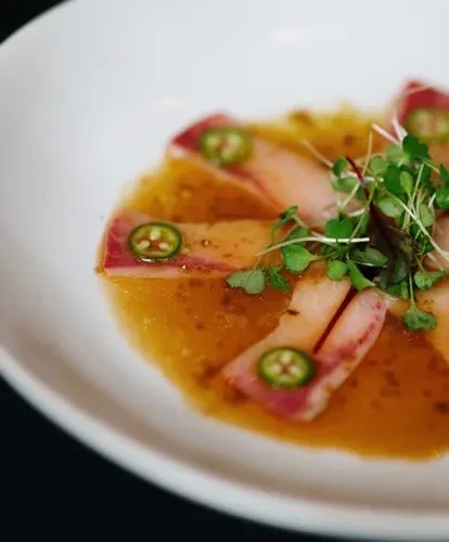 Pink fish crudo with jalapeño, microgreens, and golden sauce in a white bowl.