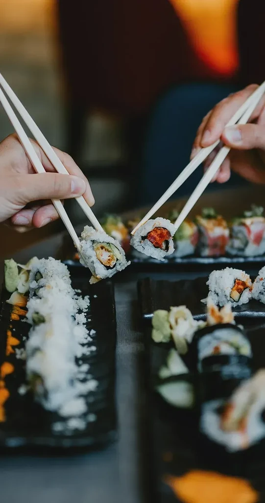 Two hands use chopsticks to pick up tempura and spicy tuna sushi.