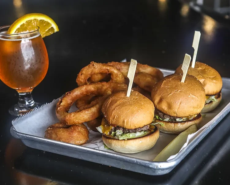 Sliders, onion rings, and beer with an orange slice on a tray.