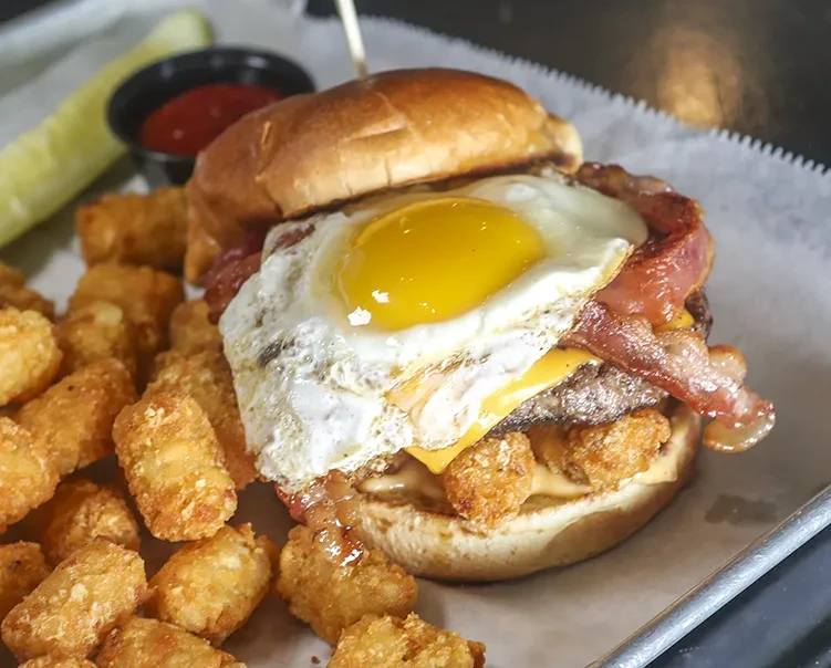 Close-up gourmet burger with fried egg, bacon, cheese, and tater tots.