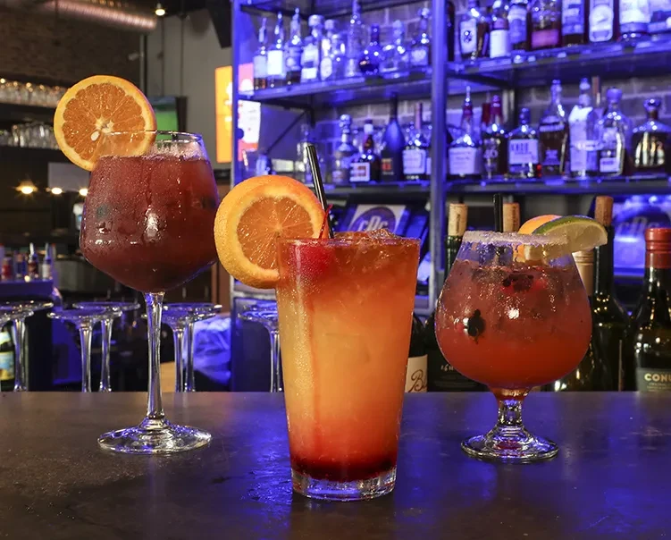 Three vibrantly colored cocktails with garnishes on a dark bar counter.