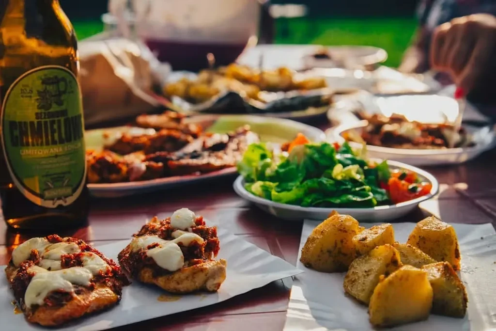 Outdoor table with grilled chicken, potatoes, salad, beer, and a person's hand.